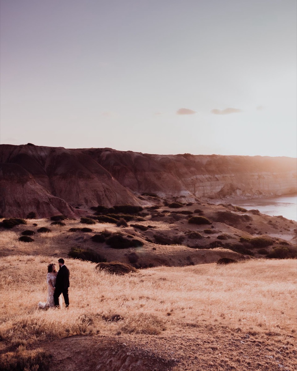 Two people at the edge of a golden-hour coastline, small against the cliffs and the sea.